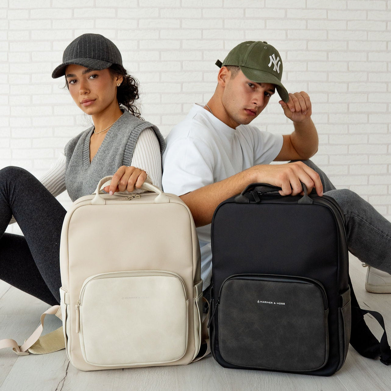 Two people holding a beige and a black backpack against a white brick wall.
