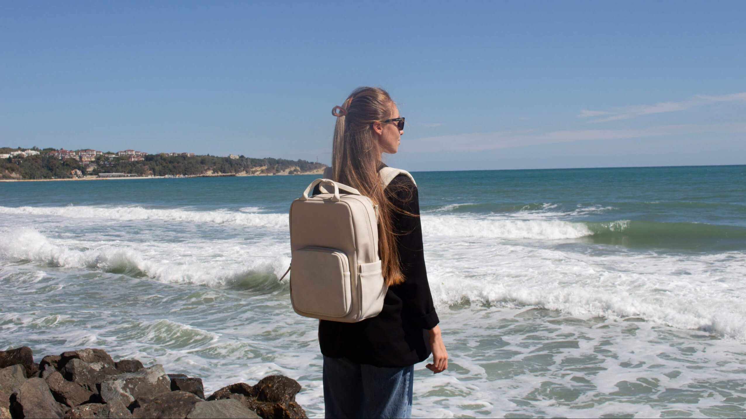 Person with a beige backpack standing on a rocky beach with ocean waves and clear sky.