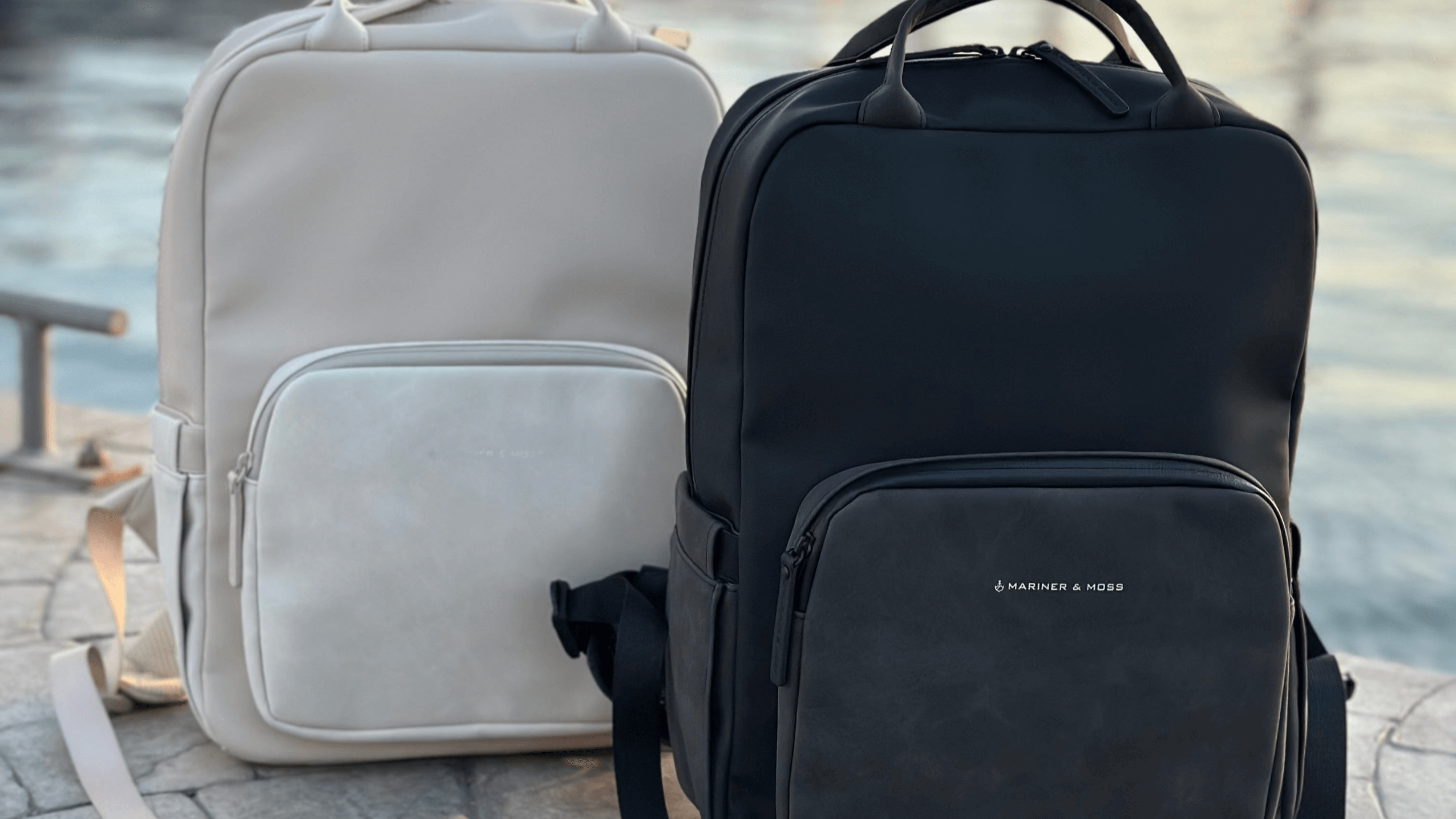 Two backpacks, one beige and one black, on a stone surface with a blurred natural background.